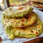 3-Ingredient Avocado Bread Recipe (Irresistibly Easy & Keto-Friendly!) 6 Golden avocado flatbreads with melted cheese and sesame seeds stacked on a parchment-lined white plate, served on a wooden board in natural light.